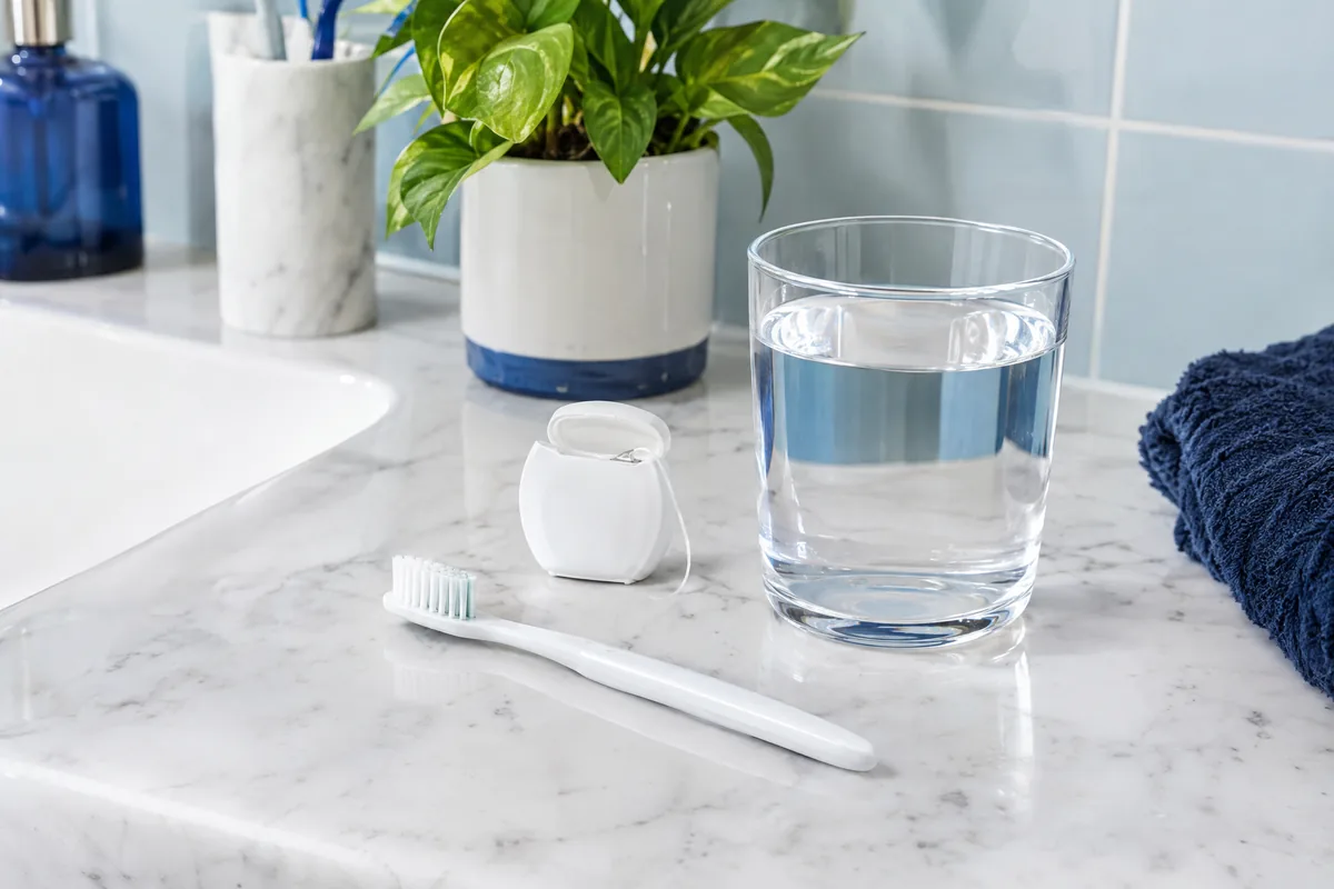 Toothbrush, dental floss, and a glass of water on a white bathroom counter in soft morning light